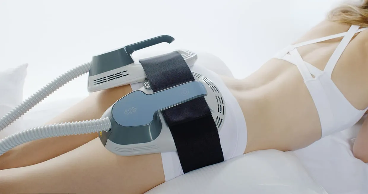 An EMSCULPT applicator strapped across a patient's abdomen with a wide black belt while she lies on the treatment bed.