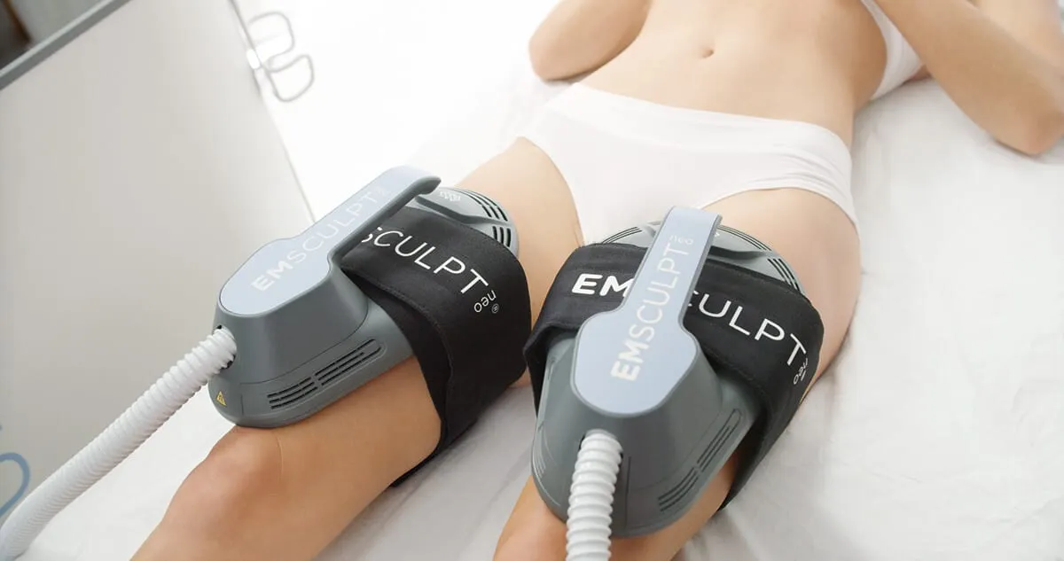 Two EMSCULPT NEO applicators belted across the fronts of both thighs while the patient lies on her back on the treatment bed.