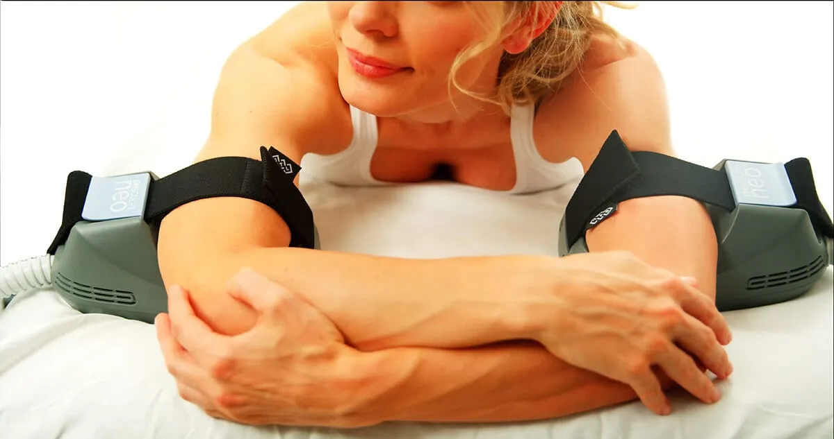 A patient lying face down with an EMSCULPT NEO applicator strapped to each upper arm, arms folded in front of her.